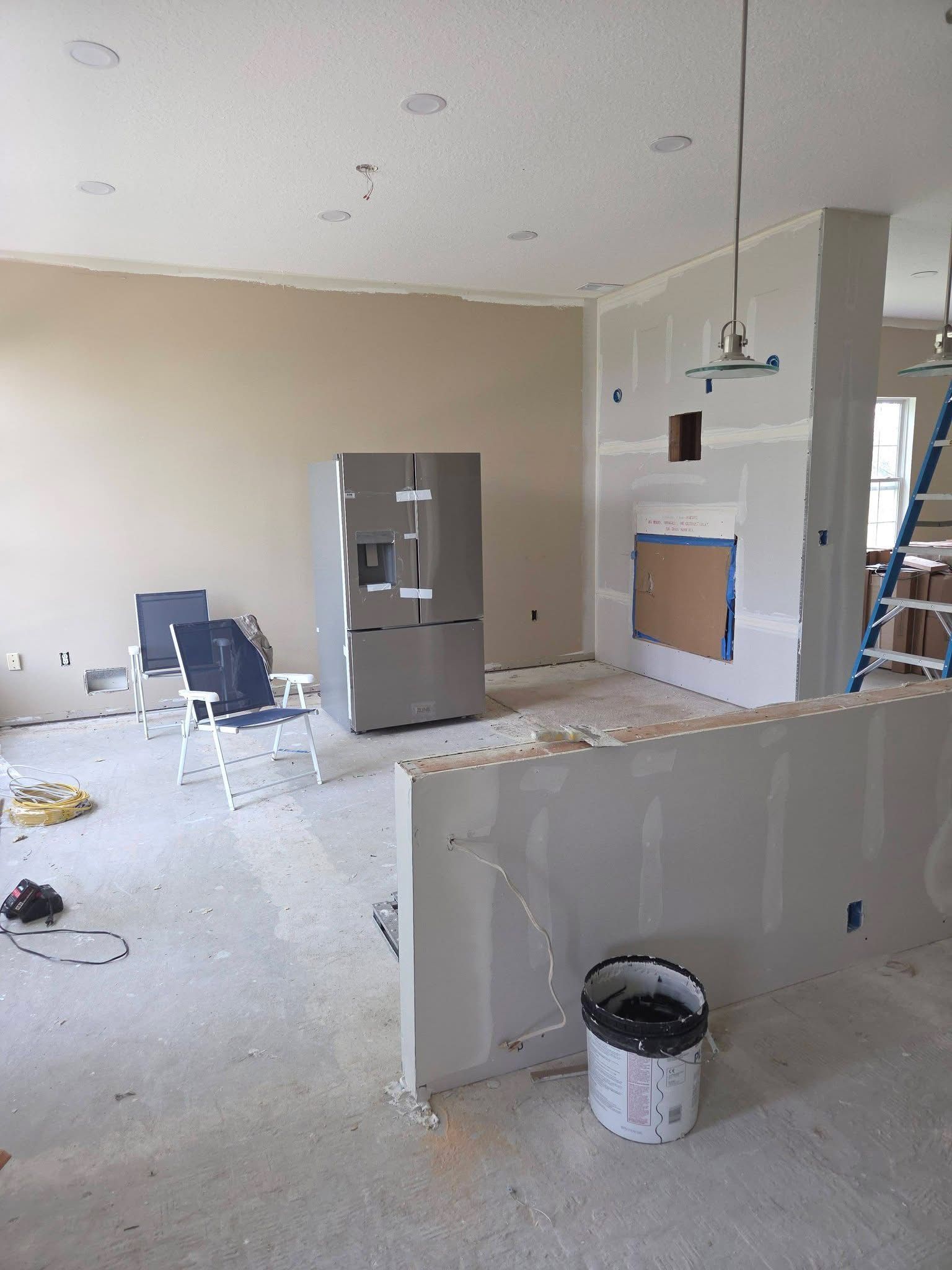 Partially finished kitchen and living room with white walls, exposed floors, and a refrigerator near the center