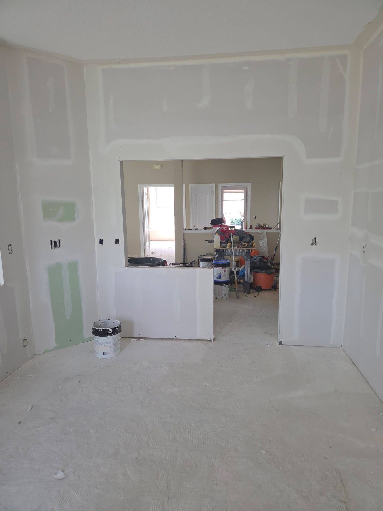 Unfinished room with drywall patches, a doorway to a cluttered room, and a white bucket on the floor.