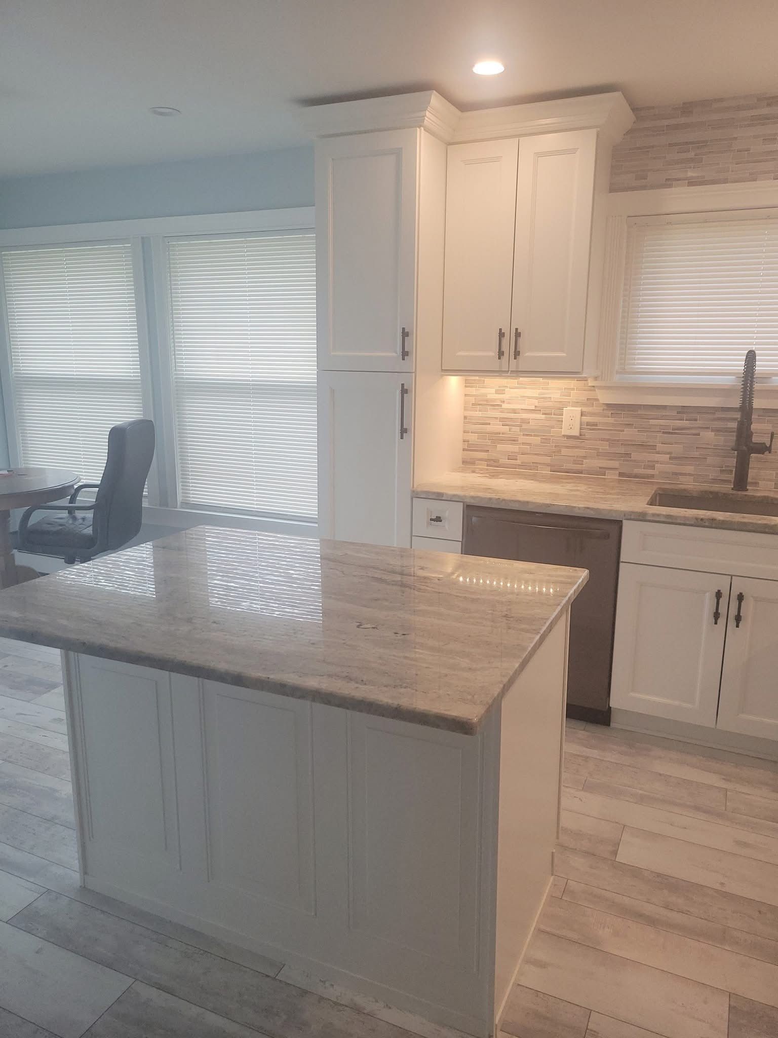 Bright white kitchen with island, marble countertops, tiled backsplash, and large windows with blinds.