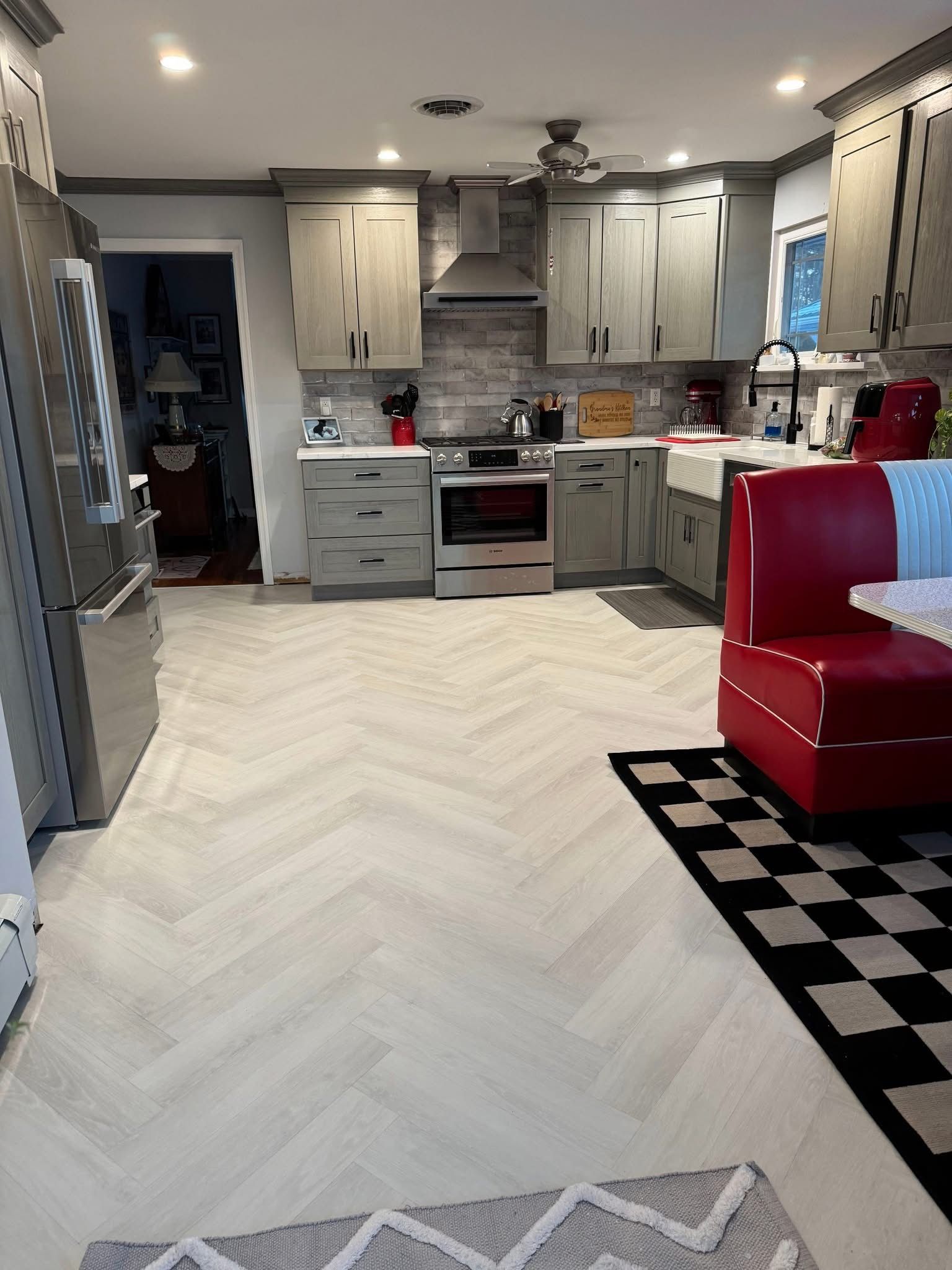Bright kitchen with gray cabinets, white herringbone floor, and red chair on a black-and-white rug.