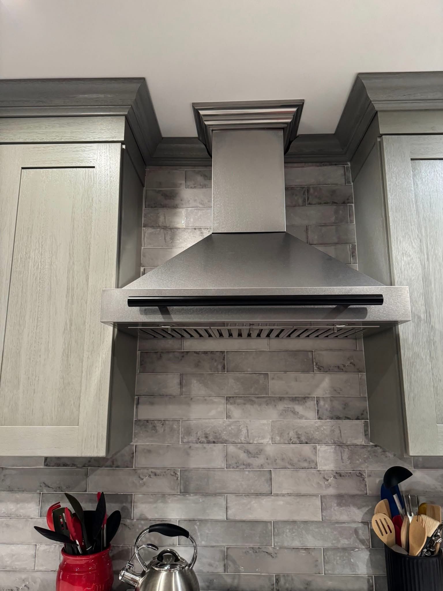 Kitchen range hood between light wood cabinets over gray tile backsplash