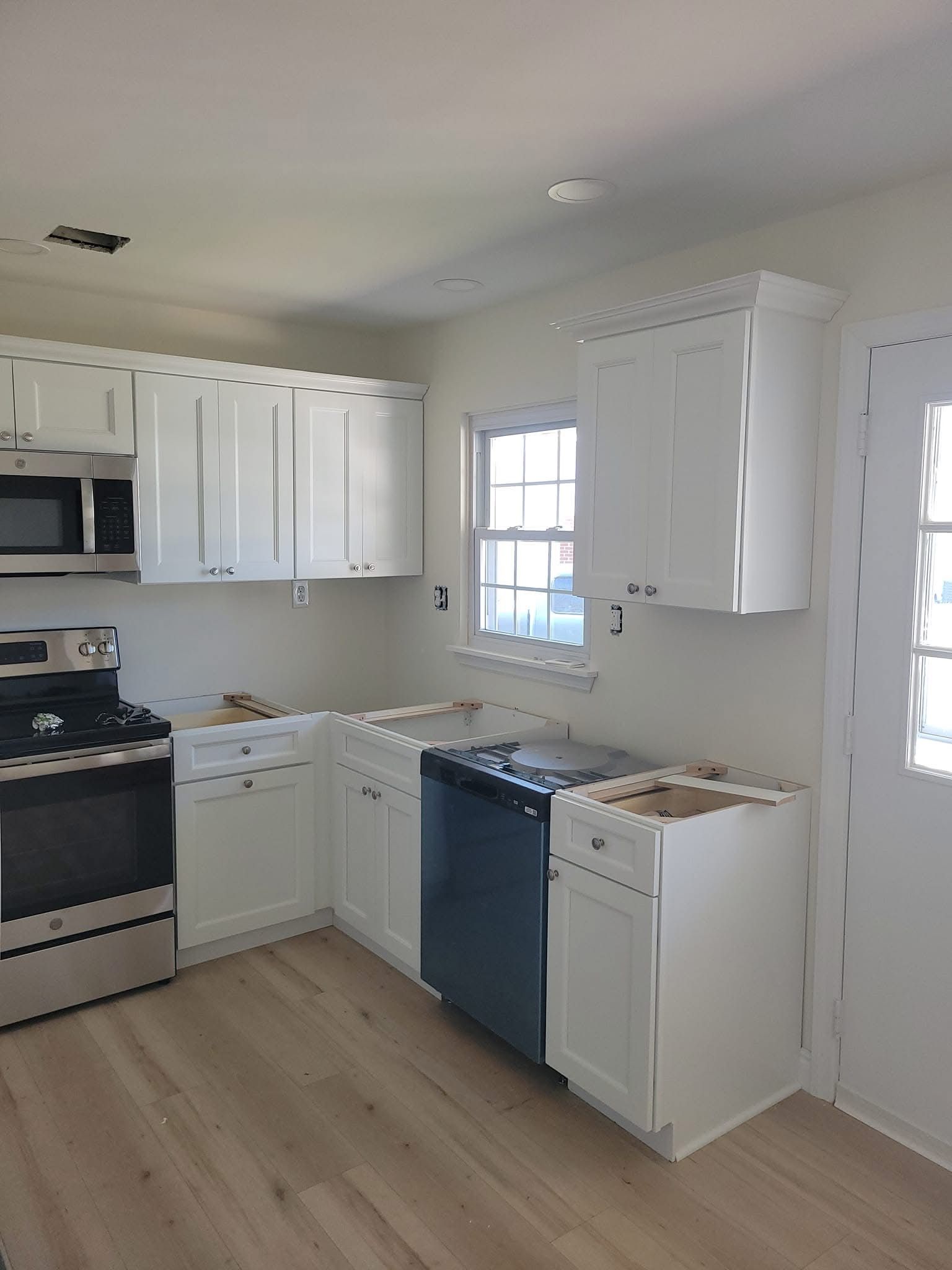 Small white kitchen with black stove, white cabinets, blue dishwasher, and light wood floors.