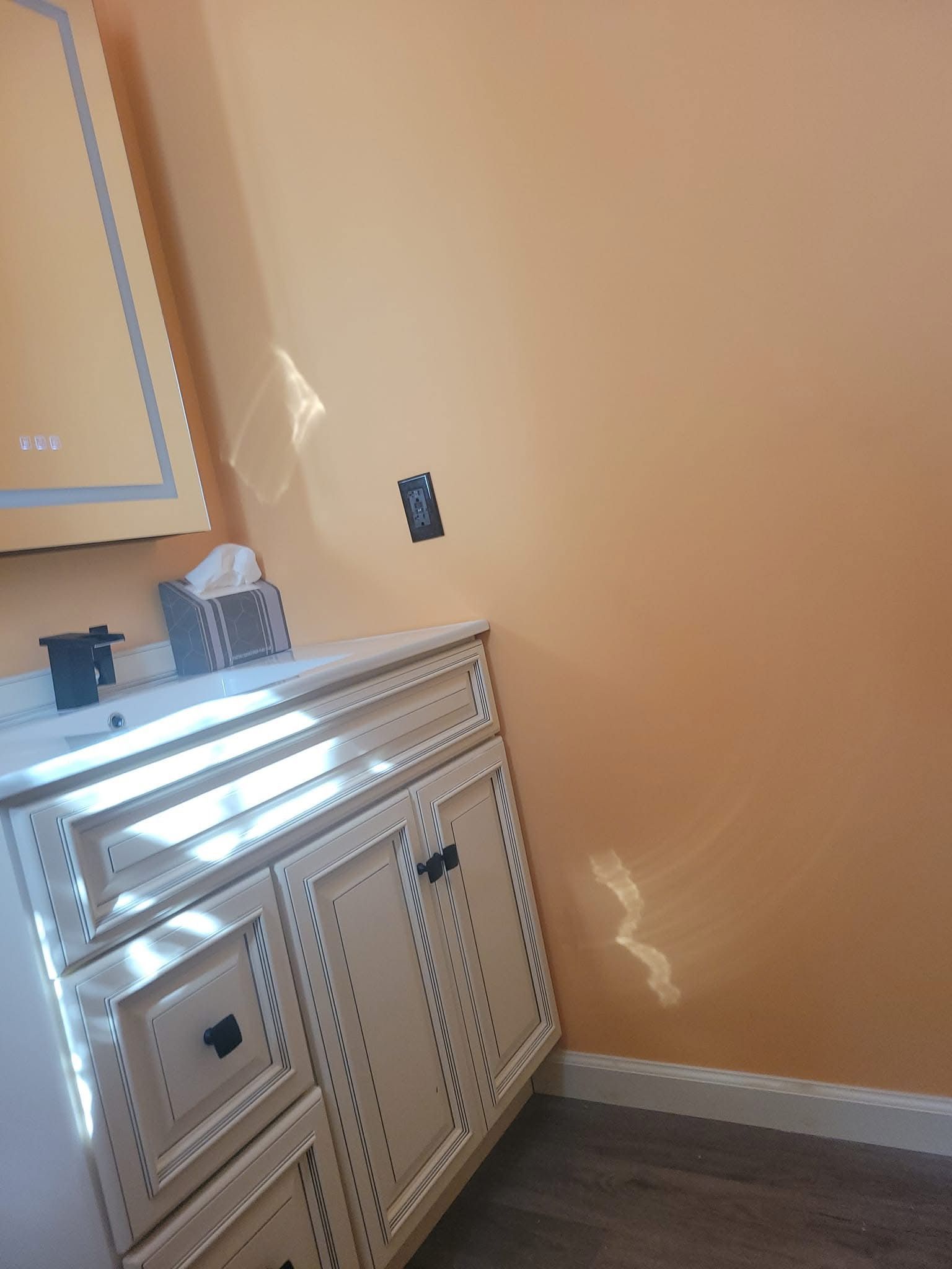 Beige bathroom with mirror, vanity, wall sconce, and sunlight on the wall.