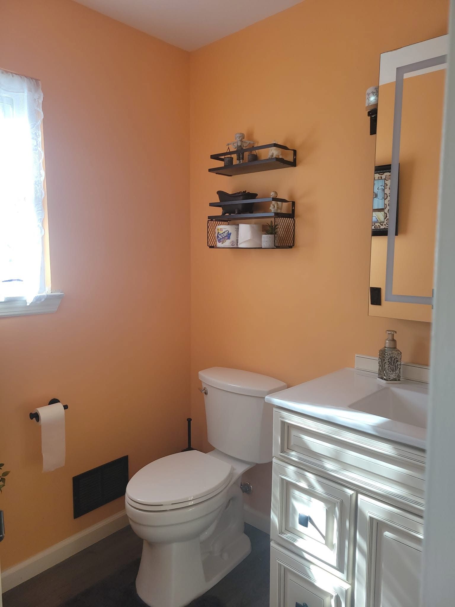 Small bathroom with peach walls, white toilet, vanity, and wall shelves beside a window.