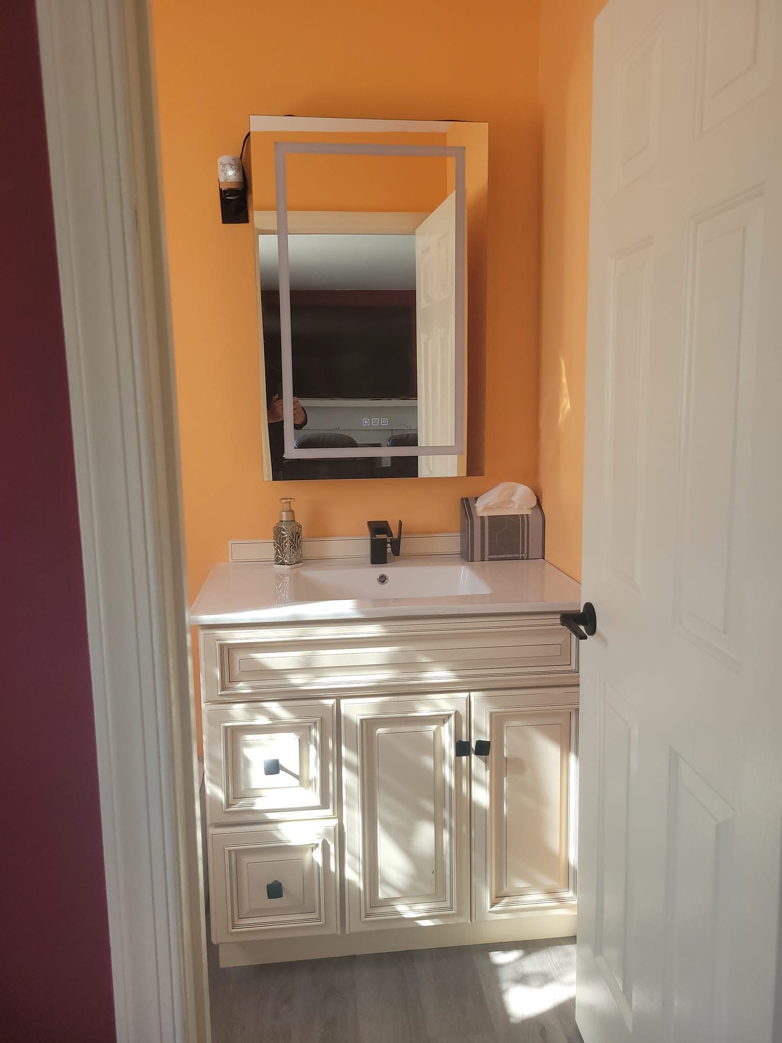 Small bathroom vanity with mirror, white cabinets, and an orange wall beside a white door.