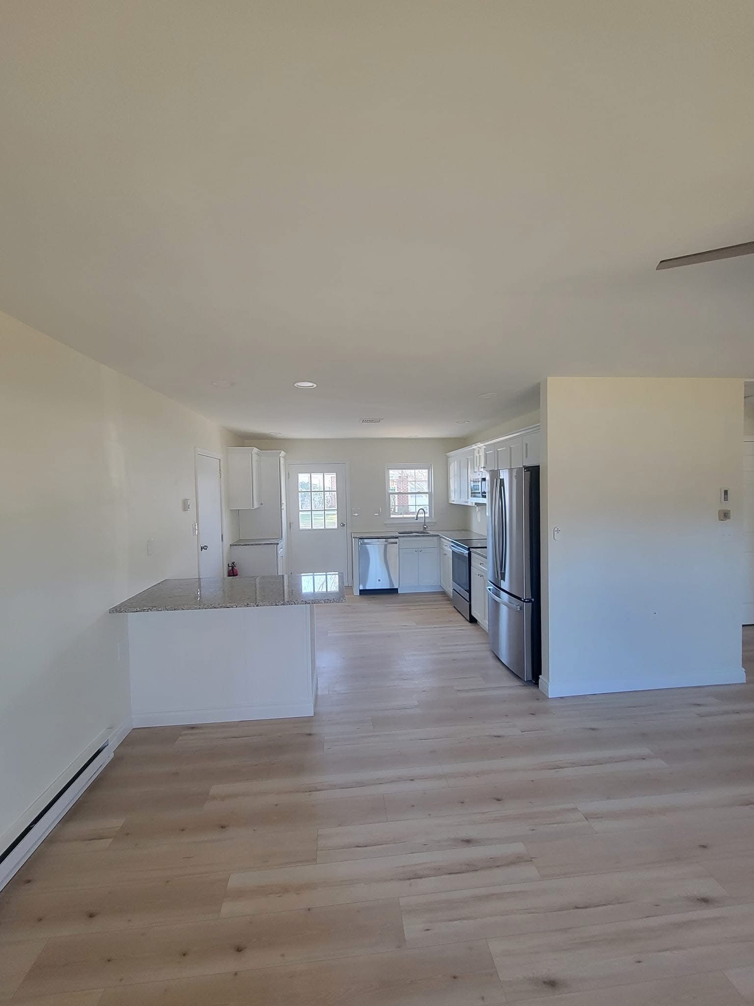 Empty modern kitchen and living area with light wood floors, white cabinets, and stainless steel fridge.