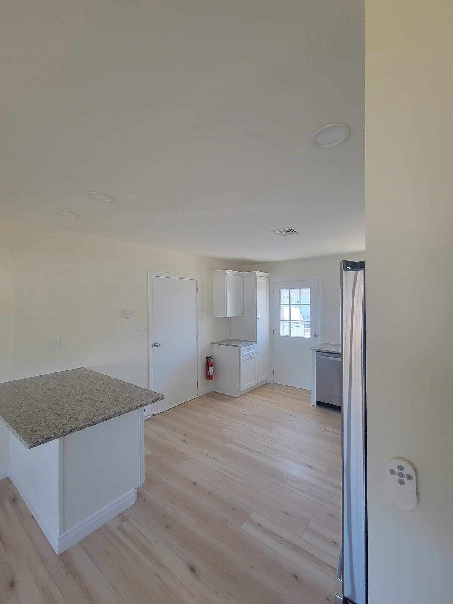 Bright empty kitchen with white cabinets, granite island, light wood floor, and a window.