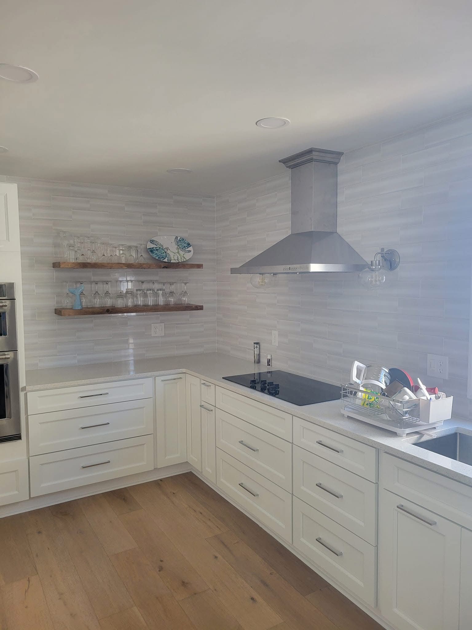 Bright white modern kitchen with wood floors, long counter, open shelves, and stainless range hood