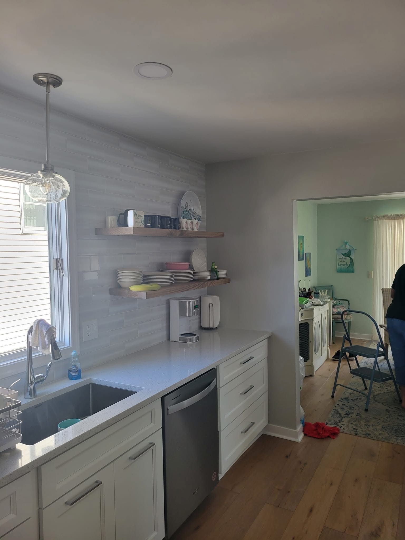 Bright kitchen with white cabinets, stainless dishwasher, open shelves, and a view into a living room.