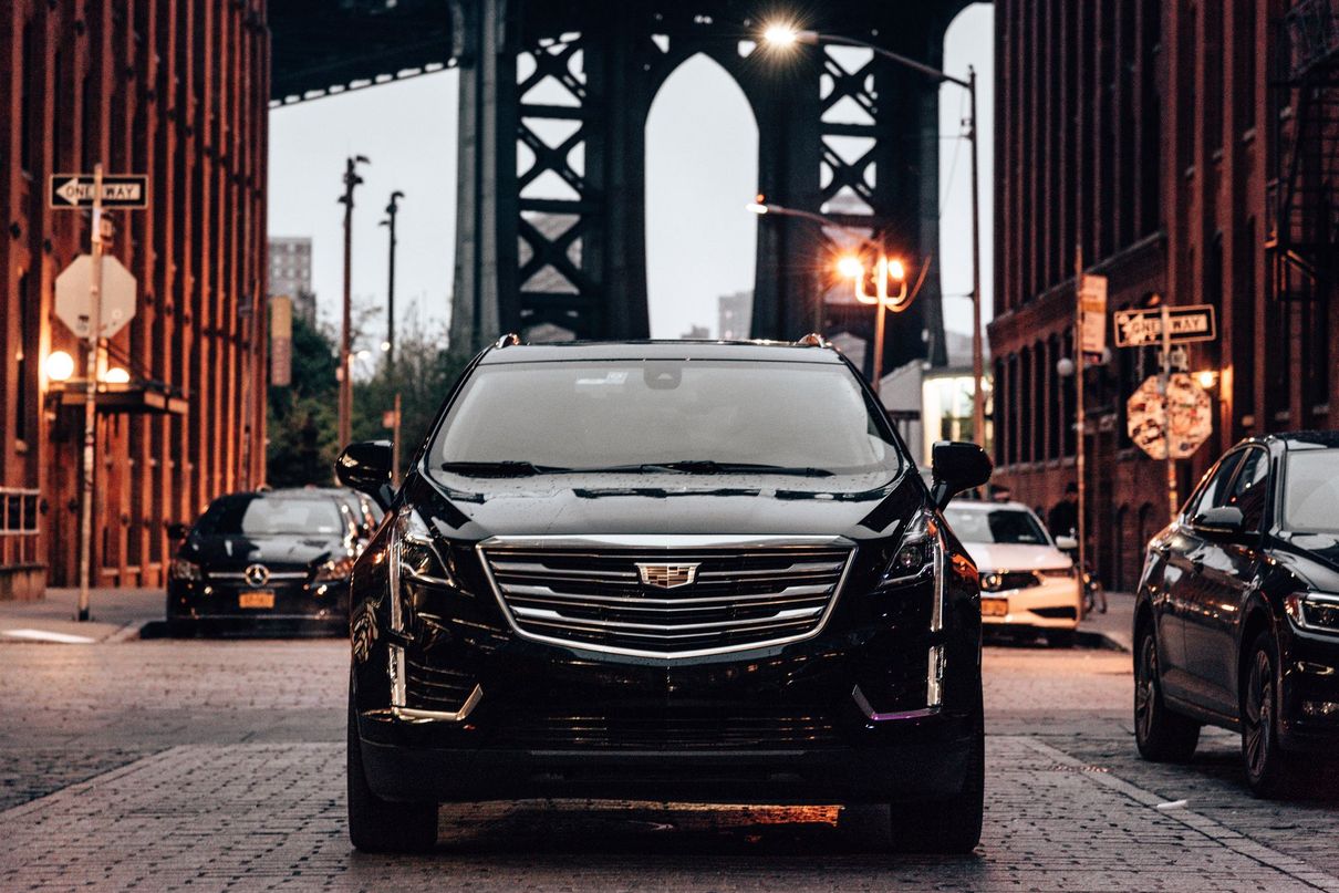 Black Cadillac SUV on a cobblestone street beneath a bridge at dusk, with city lights in the background