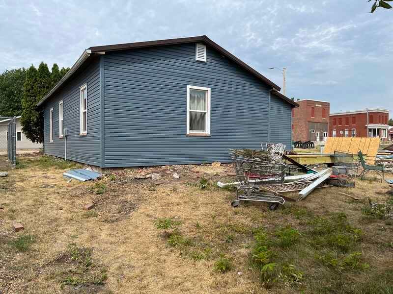 Blue house with white trim on a dirt lot, with construction debris nearby and buildings in the background