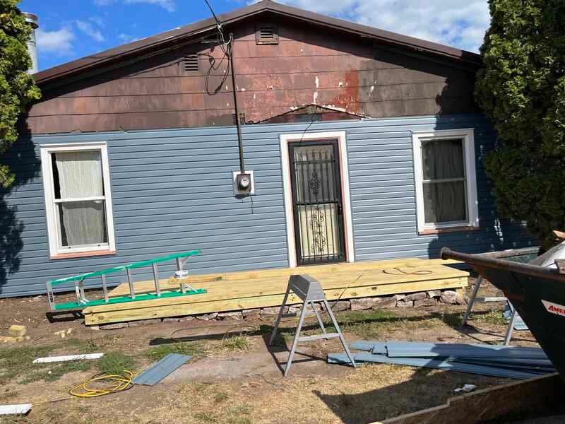 Damaged blue house exterior with broken porch boards, debris, and a ladder in front