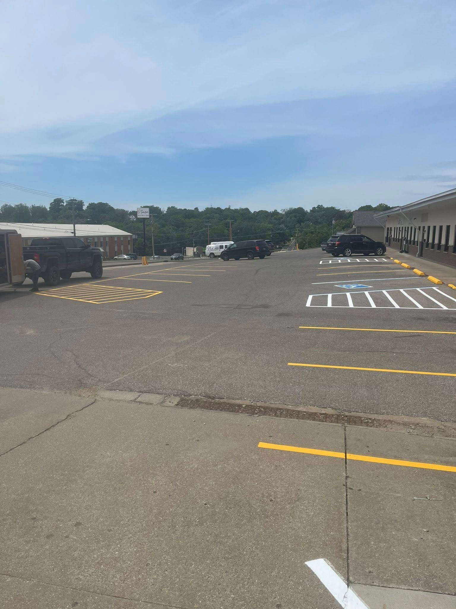 Empty parking lot with yellow lines, a few parked cars, and trees under a blue sky