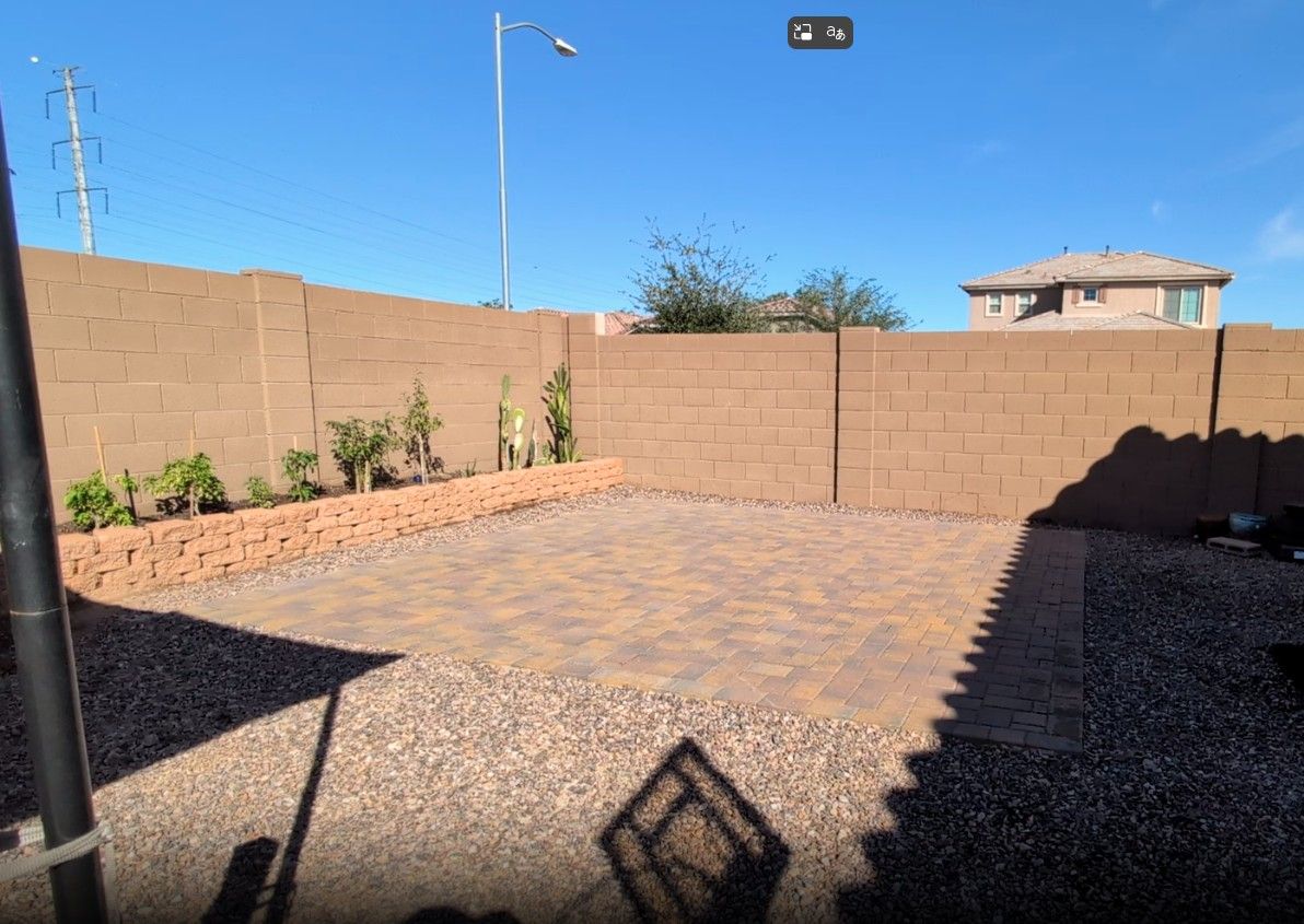 A sunny empty backyard with brick pavers, gravel borders, tan walls, and a small raised planter bed.
