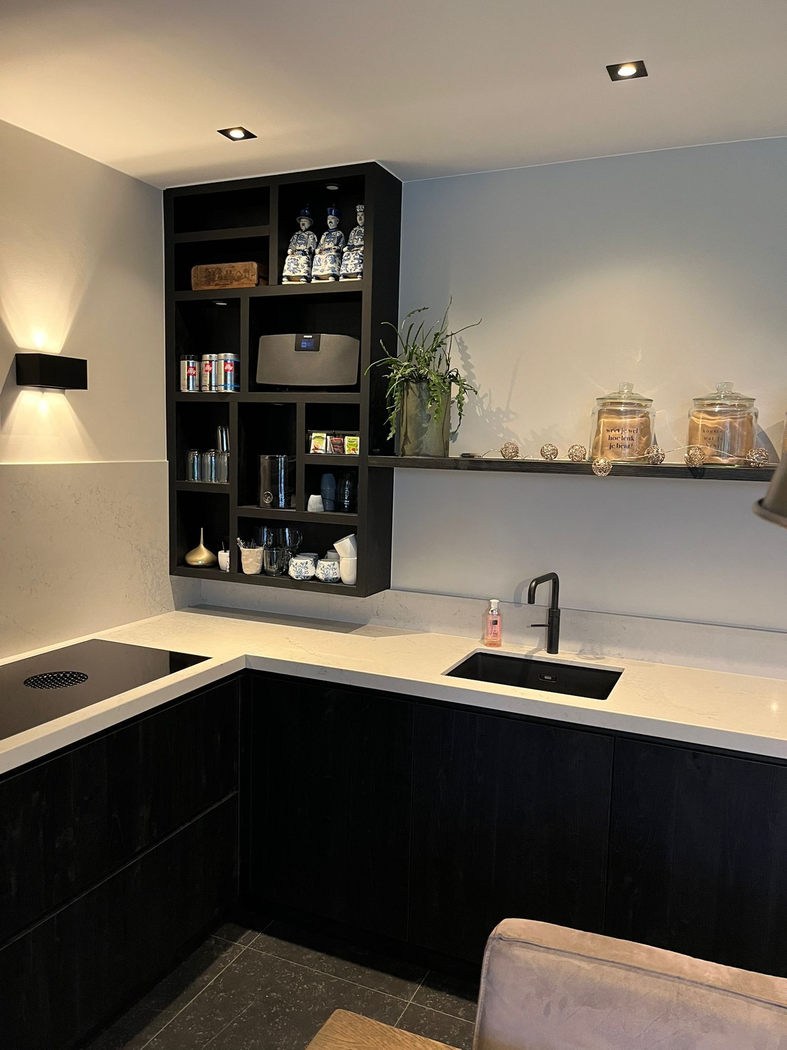 Moderne keuken met donkere kasten, ingebouwde planken en lichte werkbladen. Er is een zichtbare spoelbak.