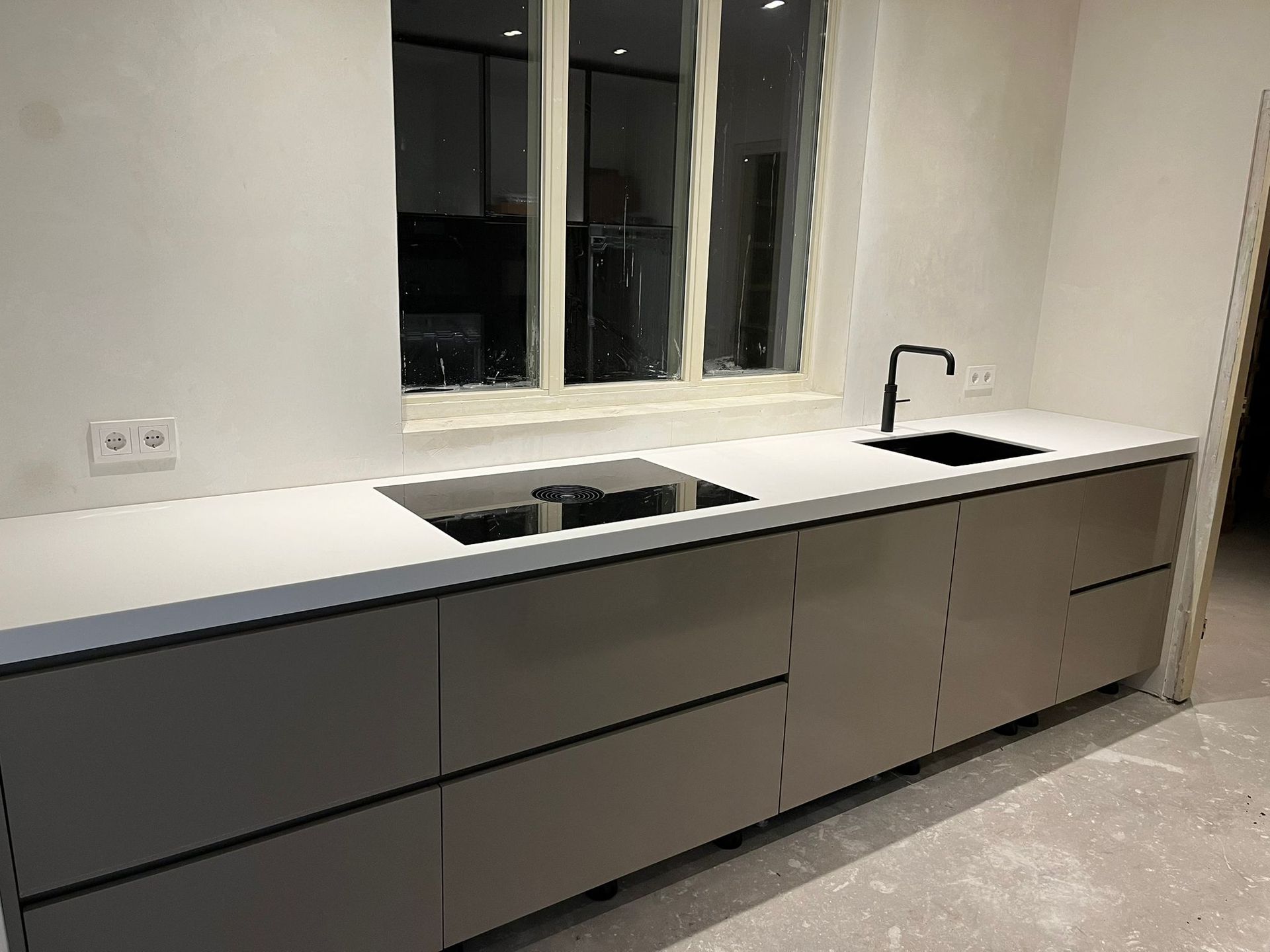 Moderne keuken met grijze kasten, wit aanrechtblad, zwarte spoelbak en raam.