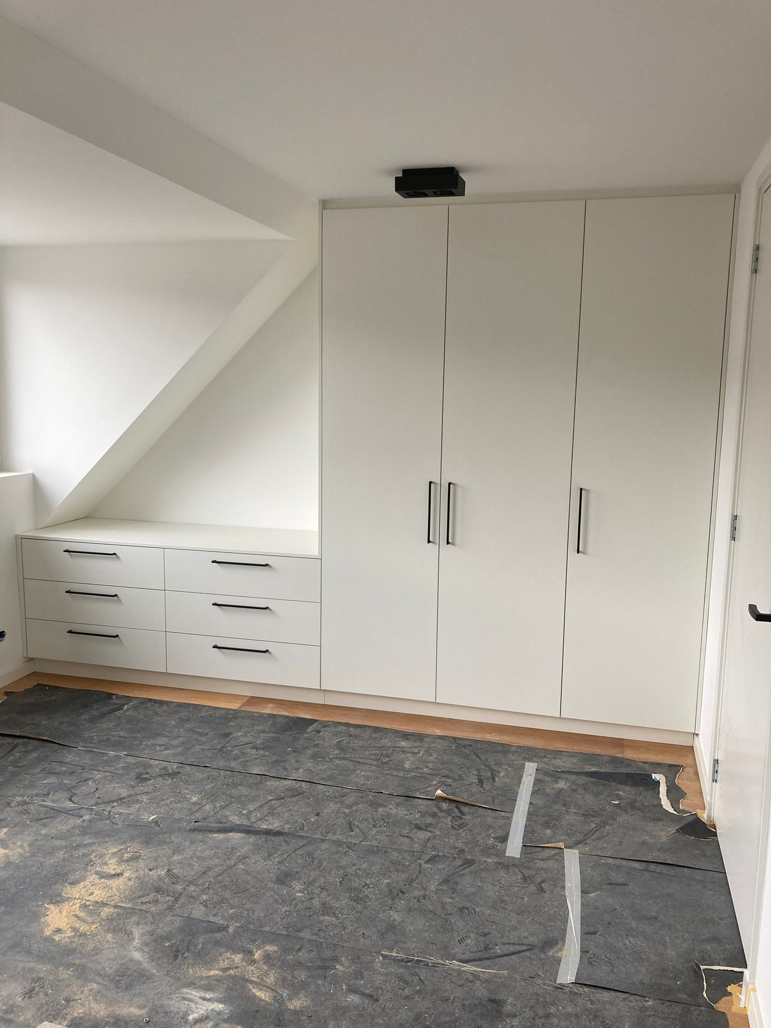 Witte kast en lades in een kamer met een schuin plafond en donkere vloer.