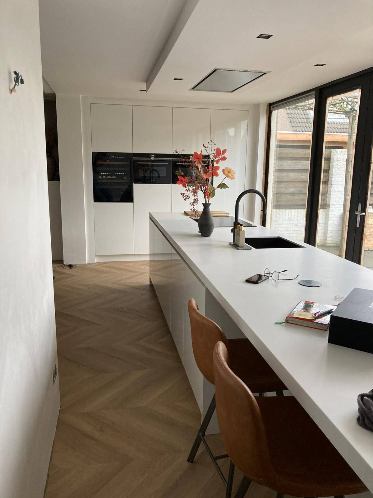 Moderne keuken met witte kasten, kookeiland en parketvloer. Voorzien van roestvrijstalen apparatuur en bruine leren barkrukken.