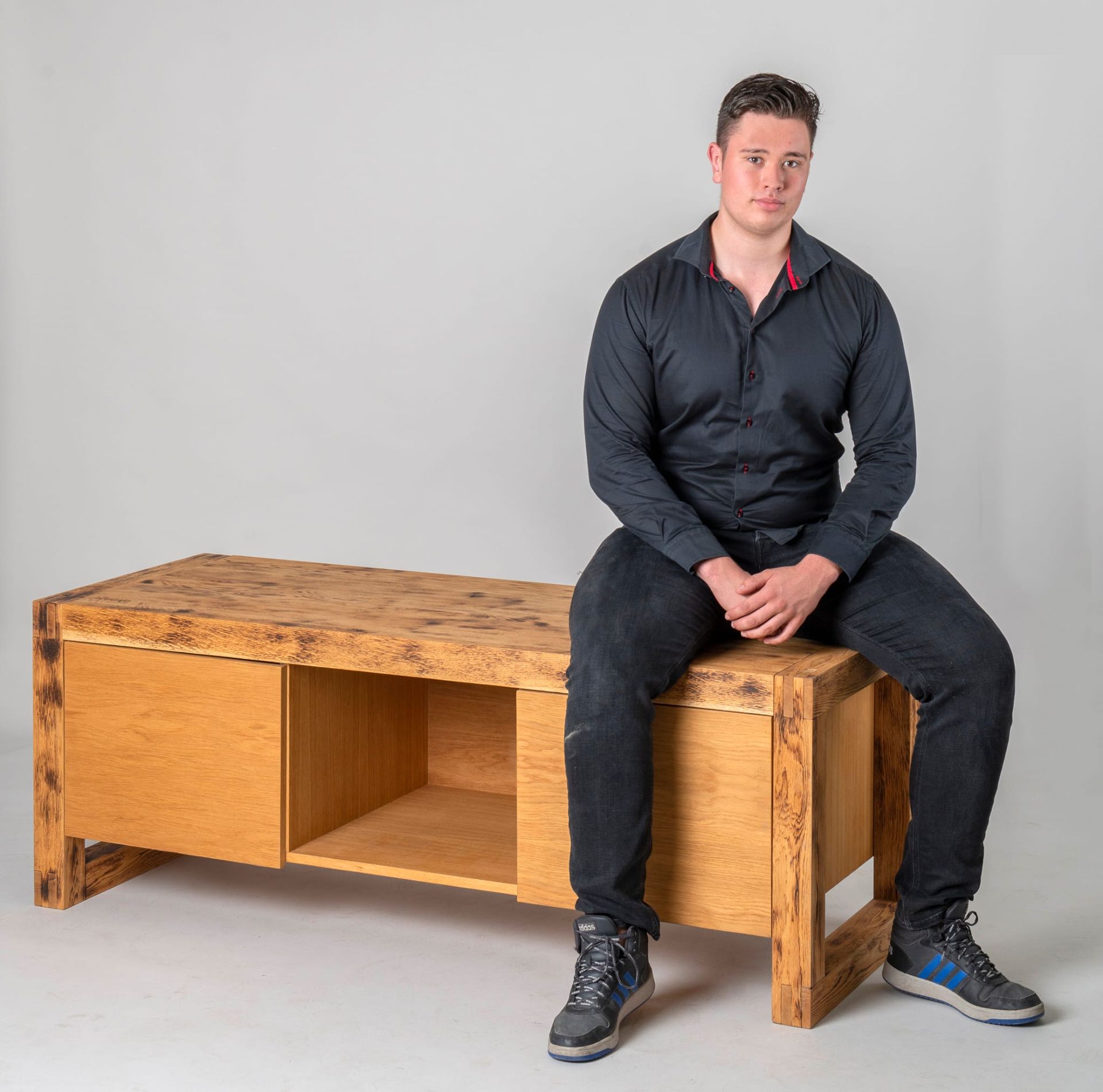 Man zittend op houten meubilair, tegen een witte achtergrond. Hij draagt ​​een donker shirt en een zwarte spijkerbroek.