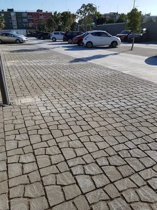 Pavimentazione in ciottoli in un parcheggio con auto parcheggiate, edifici e alberi sotto un cielo limpido.