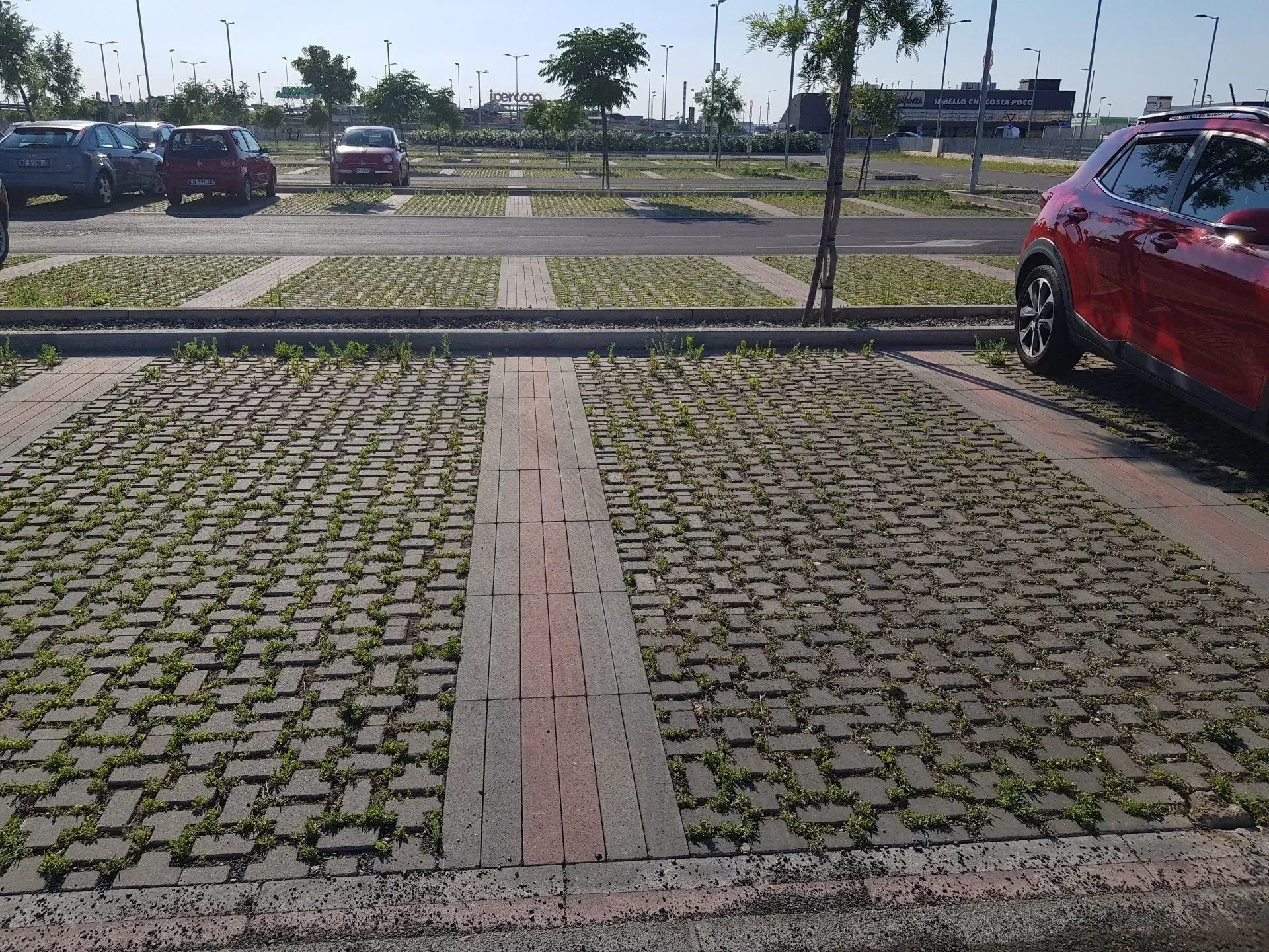 Parcheggio con pavimentazione in mattoni, alcune con erba. Un'auto rossa sulla destra, altre auto sullo sfondo. Giornata di sole.