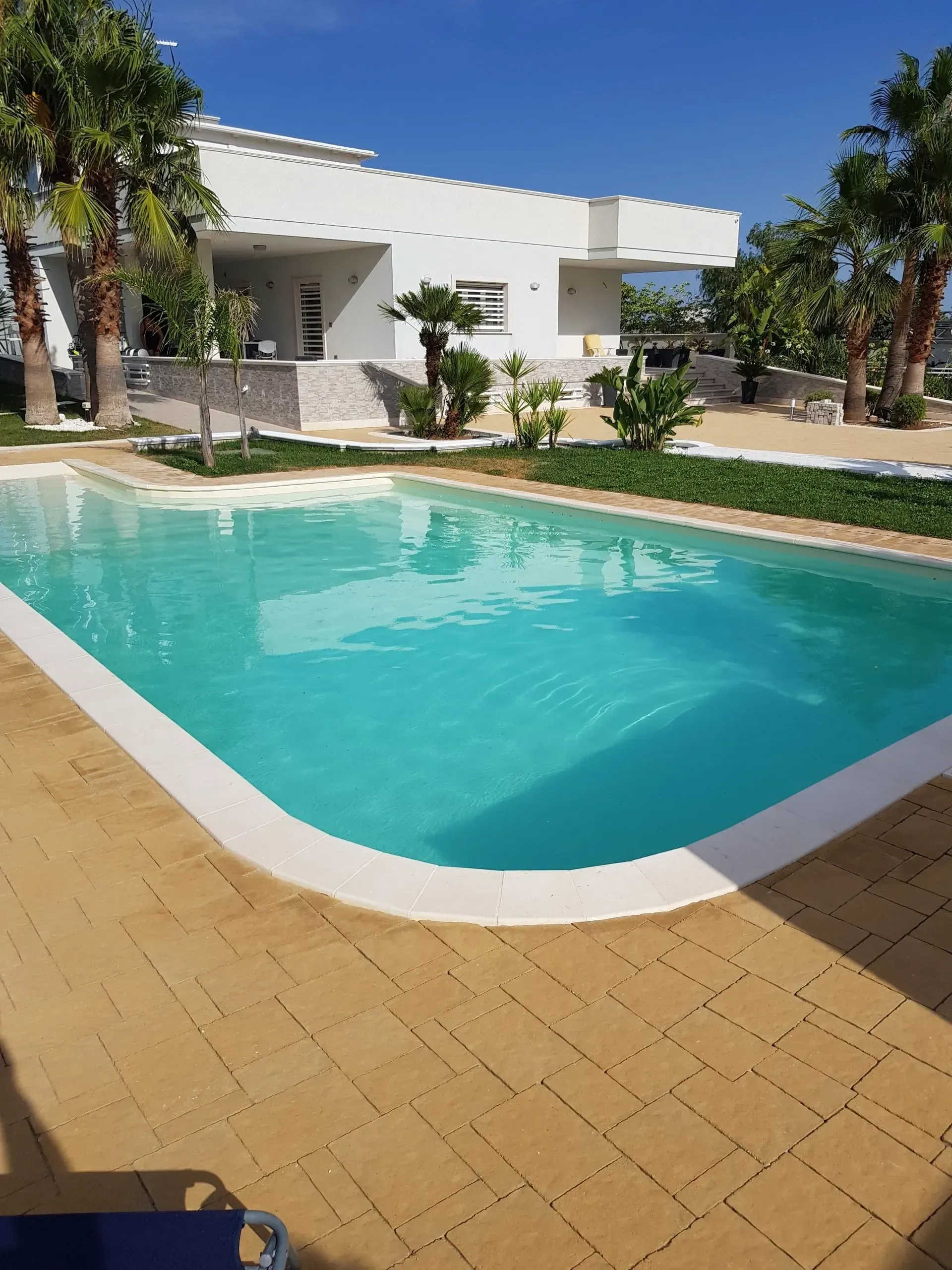 Vista a bordo piscina di una villa bianca con acqua turchese, palme e patio in pietra color sabbia sotto un cielo azzurro.