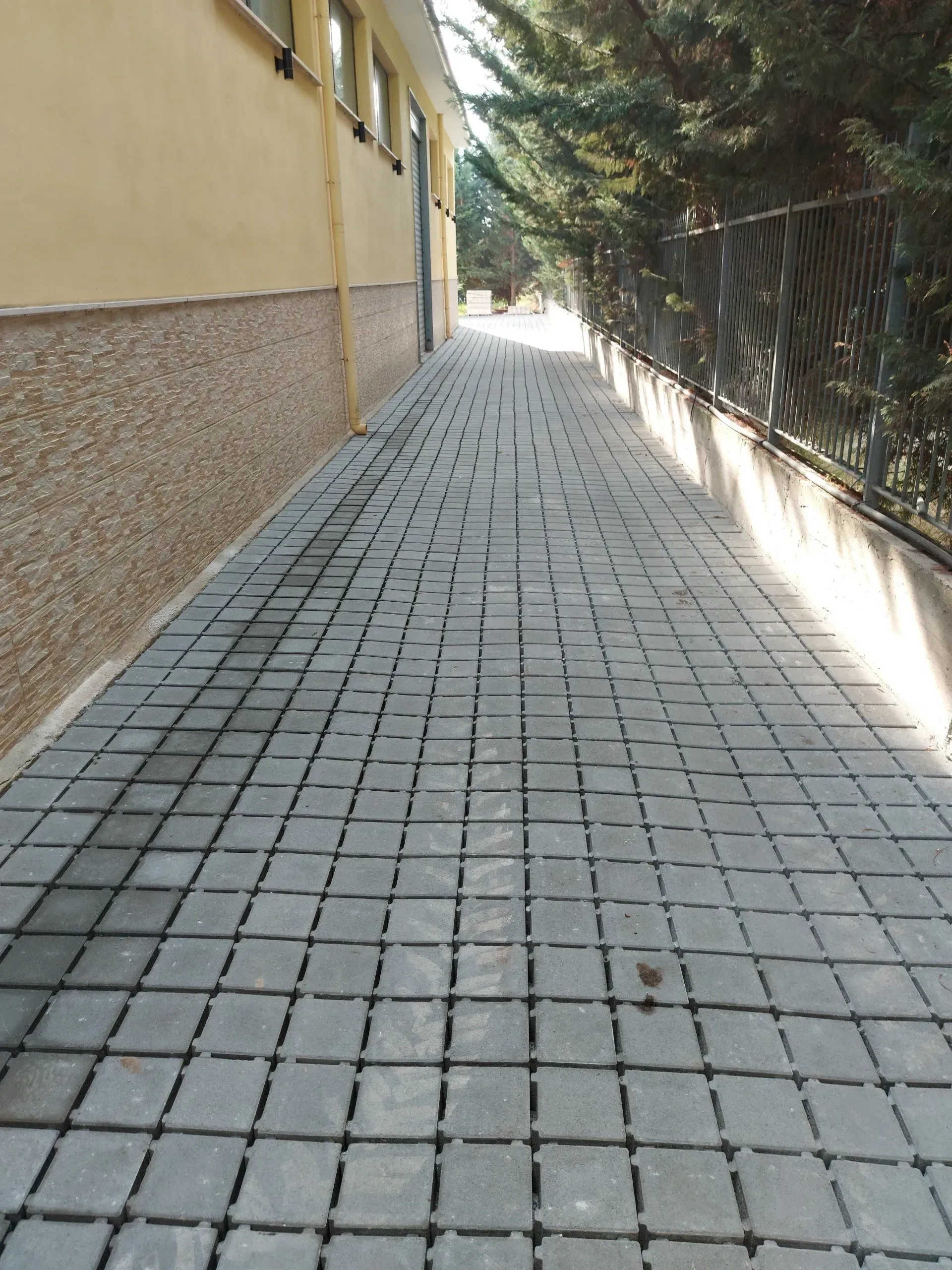 Passaggio pedonale pavimentato tra un edificio beige e una recinzione con alberi.