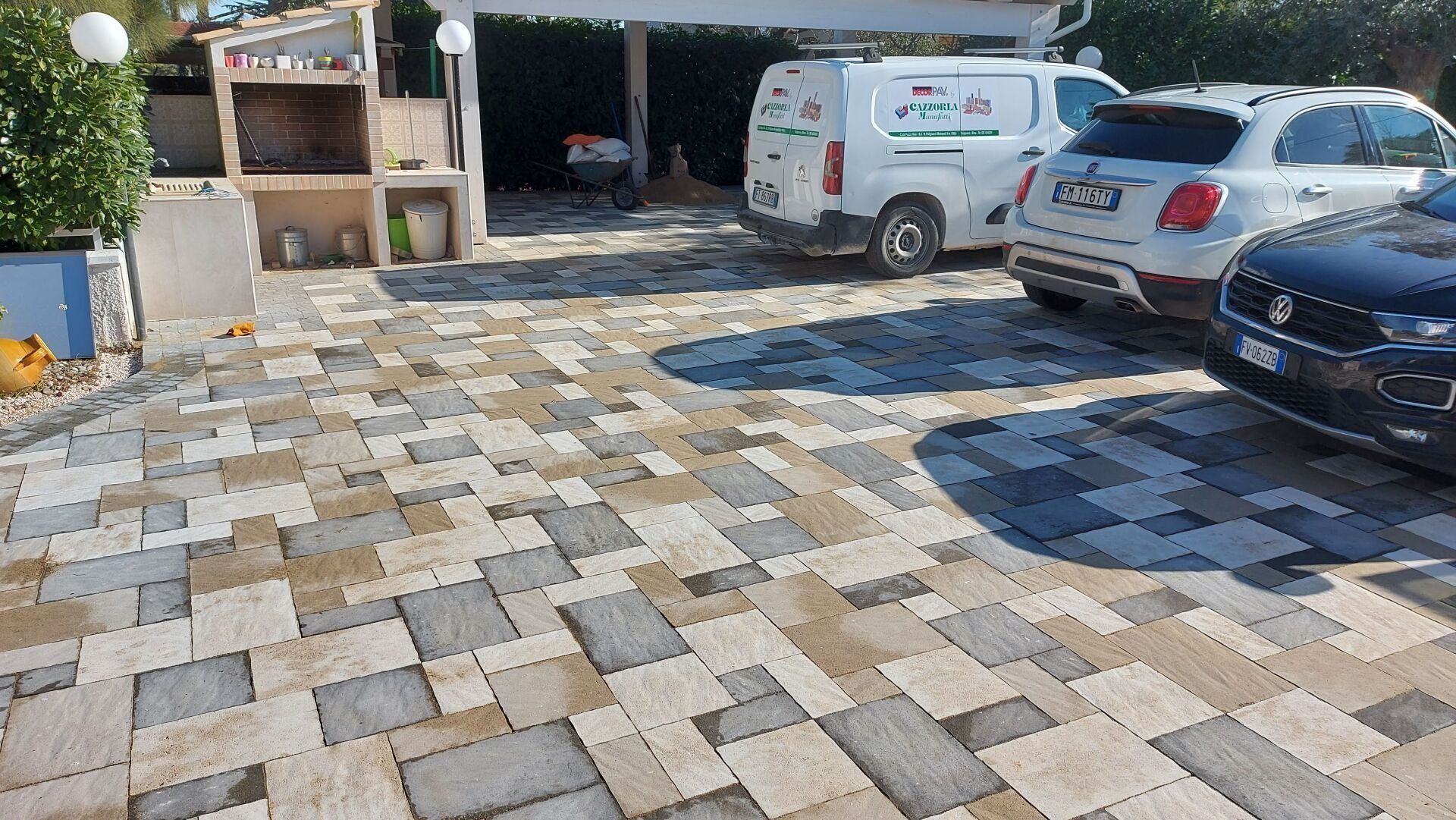 Vialetto lastricato in mattoni con veicoli parcheggiati; furgone bianco con logo, SUV bianco e nero, auto scura. Area barbecue sulla sinistra. Ambiente esterno soleggiato.
