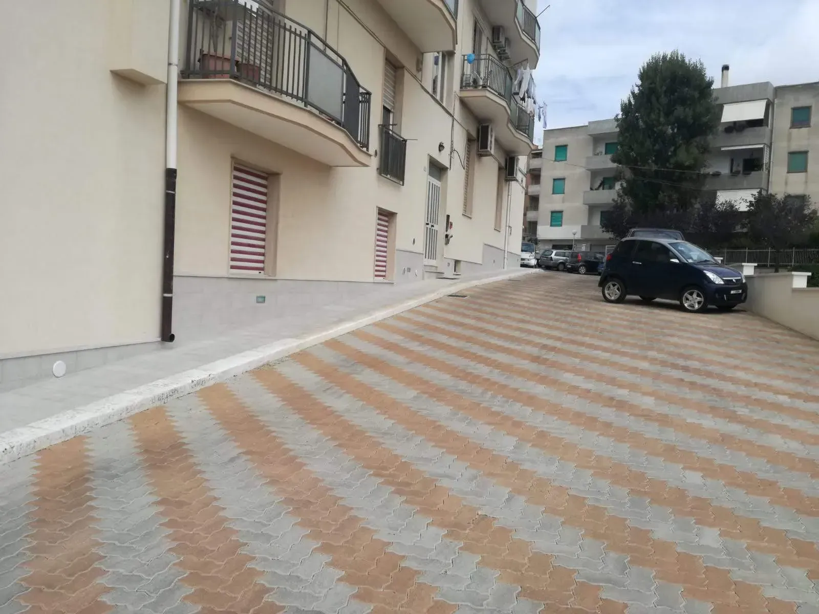 Il parcheggio pavimentato in mattoni è in salita, con auto parcheggiate e un edificio dotato di balconi e tende da sole.