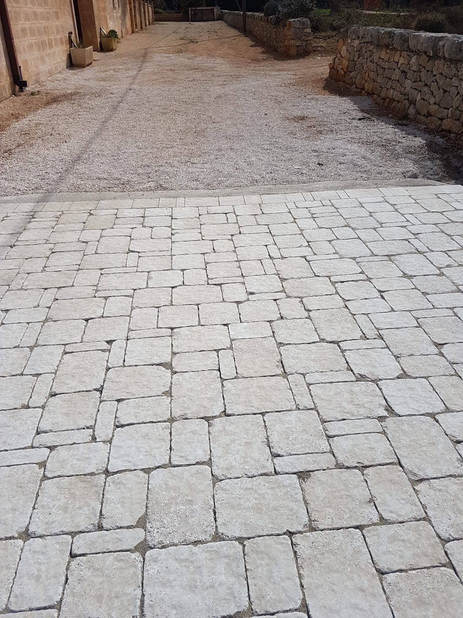 Vialetto lastricato in pietra che si trasforma in sentiero ghiaioso. Edifici e muretto visibili.