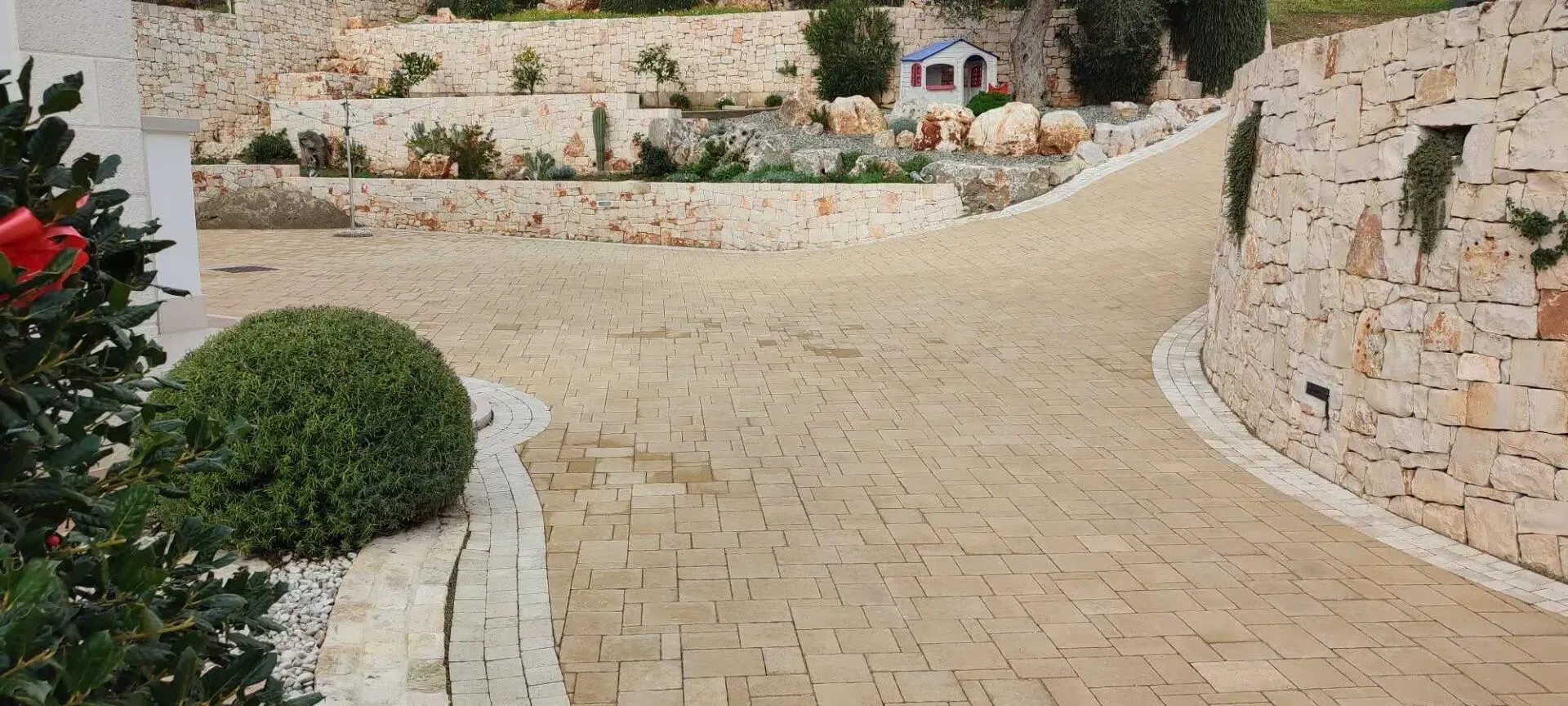 Il vialetto lastricato in pietra curva oltrepassando un cespuglio circolare e un muro di contenimento in pietra. Sullo sfondo si intravede un giardino terrazzato.