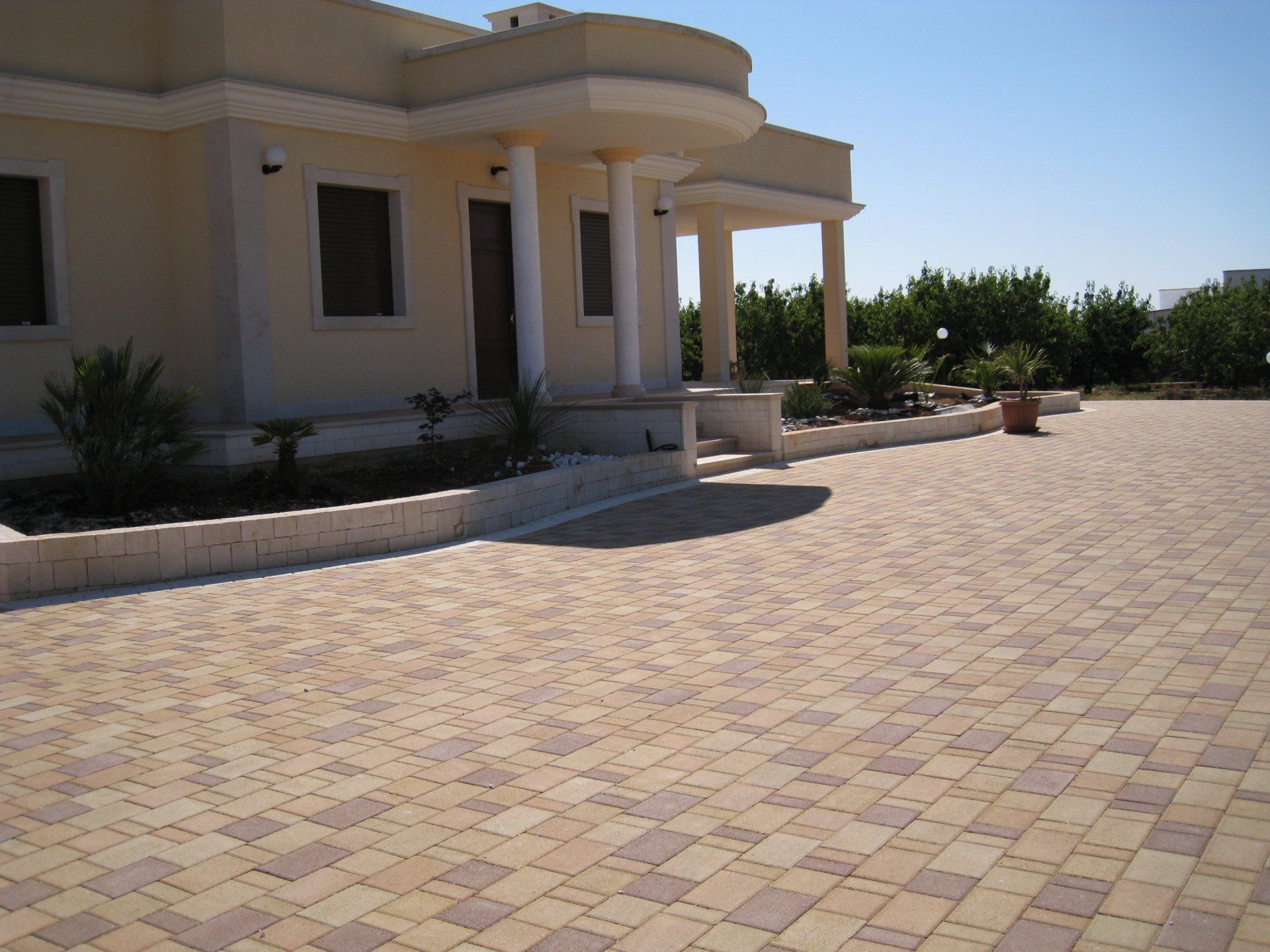 Vialetto pavimentato in mattoni color marrone chiaro e beige che conduce a un edificio chiaro con colonne, sotto un cielo azzurro e terso.