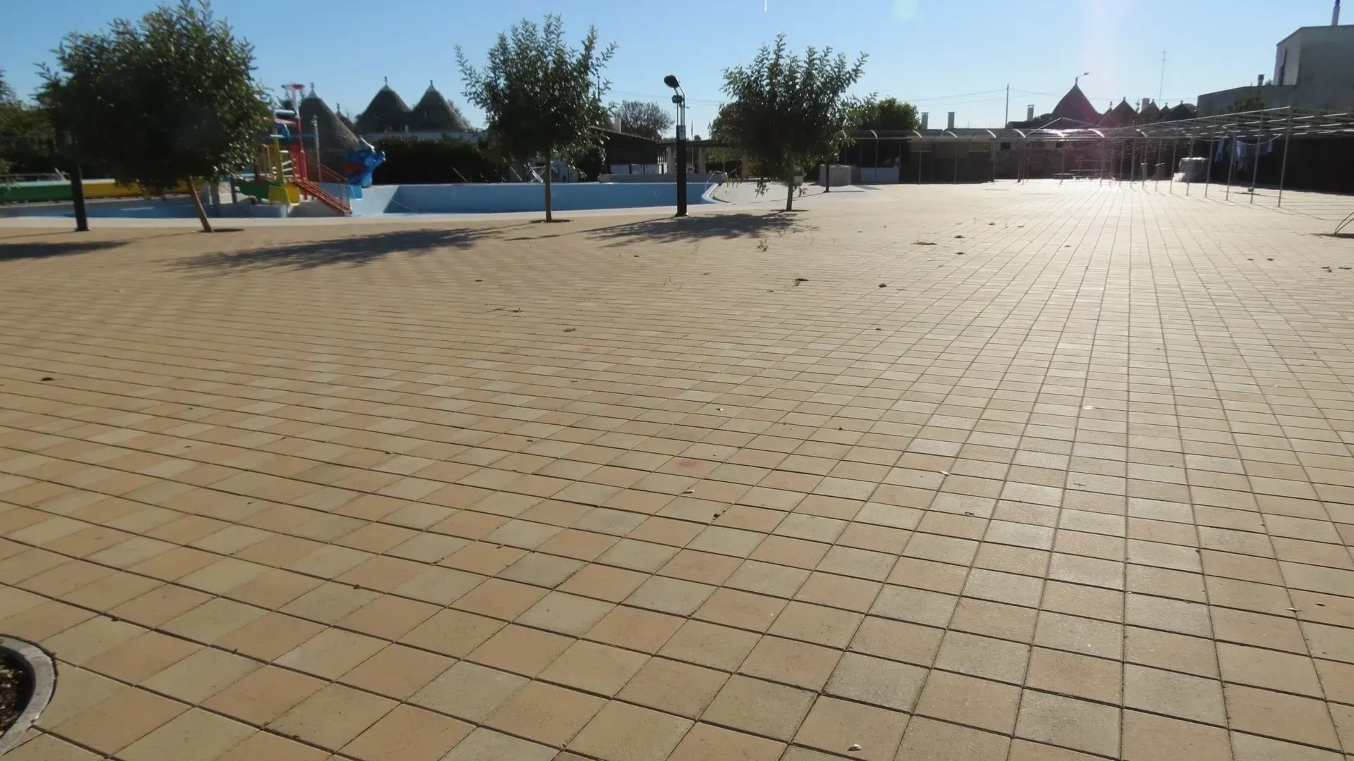 Piazza pavimentata in mattoni con piccoli alberi, con una piscina e un parco giochi sullo sfondo.