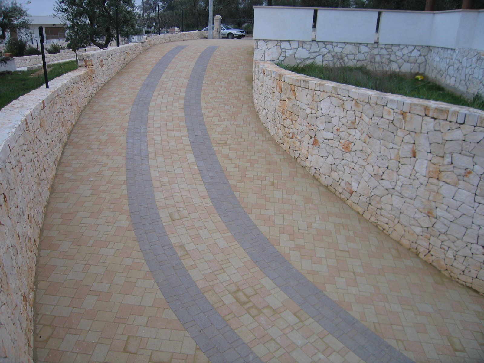 Vialetto curvilineo in pietra con tracce di pneumatici grigi, delimitato da muri in pietra.