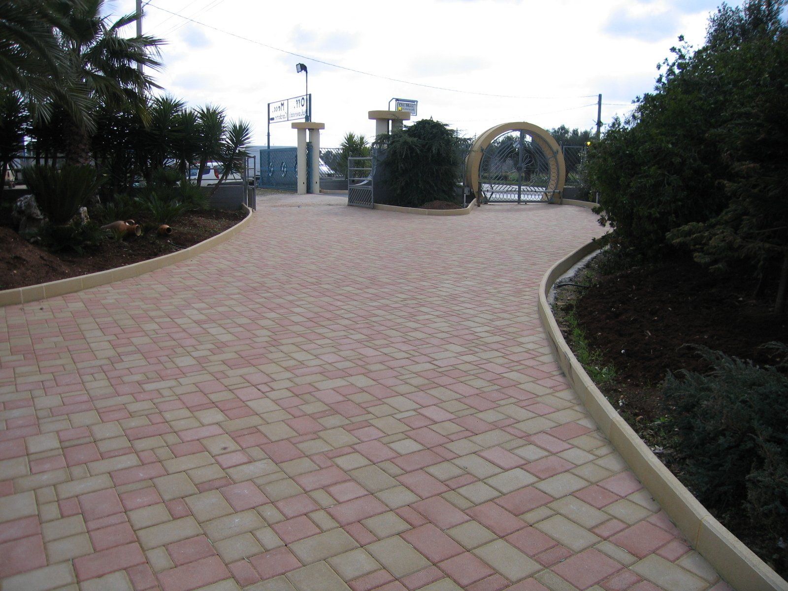 Il sentiero lastricato in mattoni curva verso un ingresso recintato, con alberi su entrambi i lati e un arco decorativo.