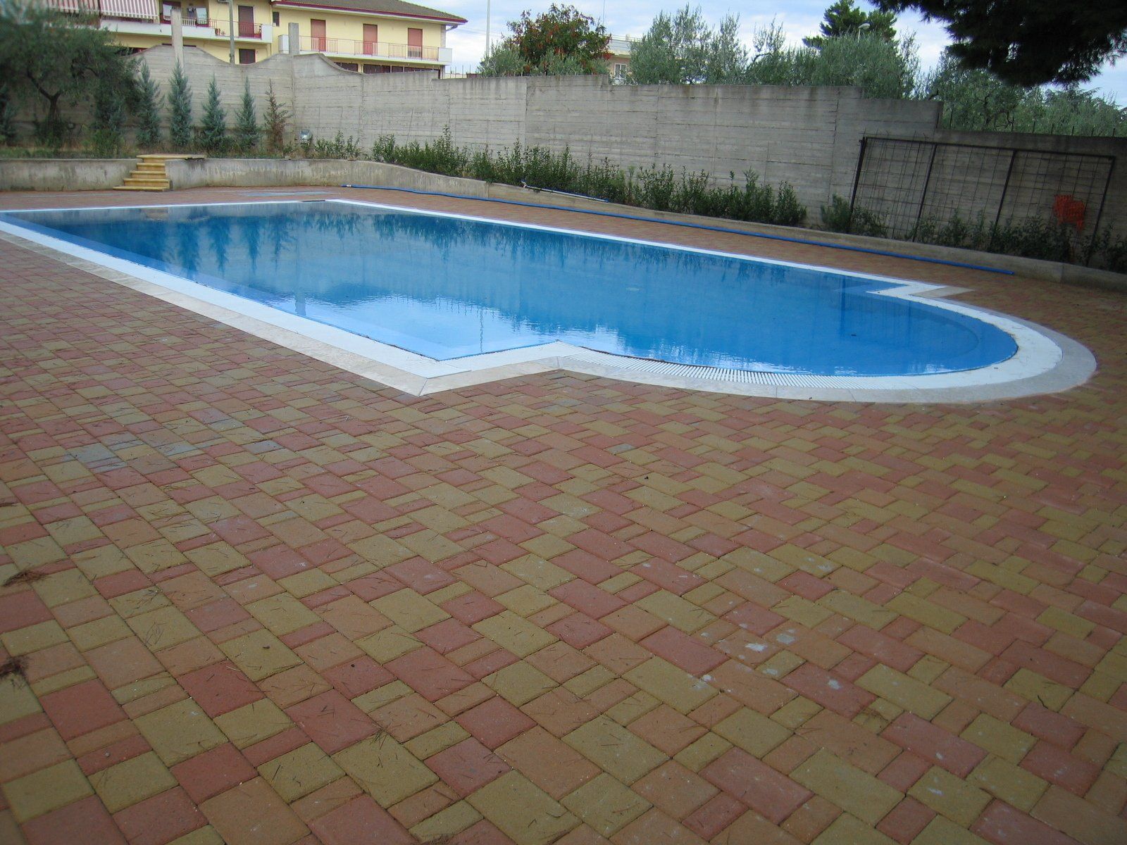 Piscina con acqua azzurra circondata da mattoni. Sullo sfondo un muro di cemento e un edificio.