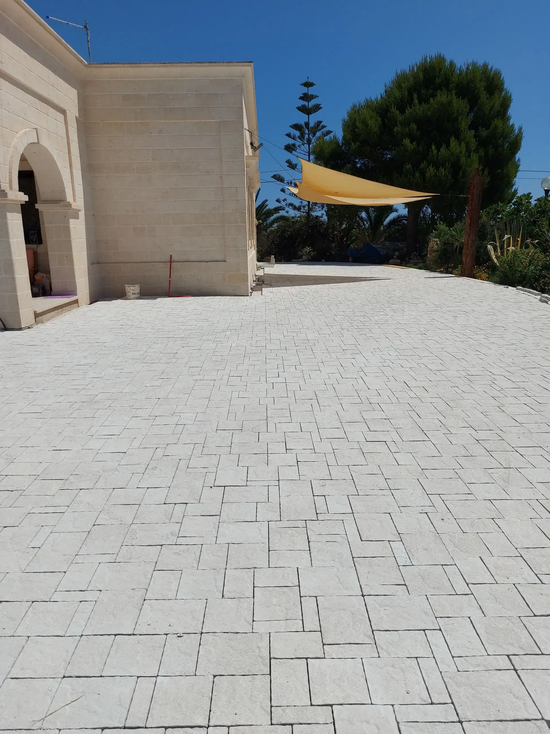 Patio esterno pavimentato in pietra bianca; edificio beige e vela ombreggiante visibili; giornata soleggiata.