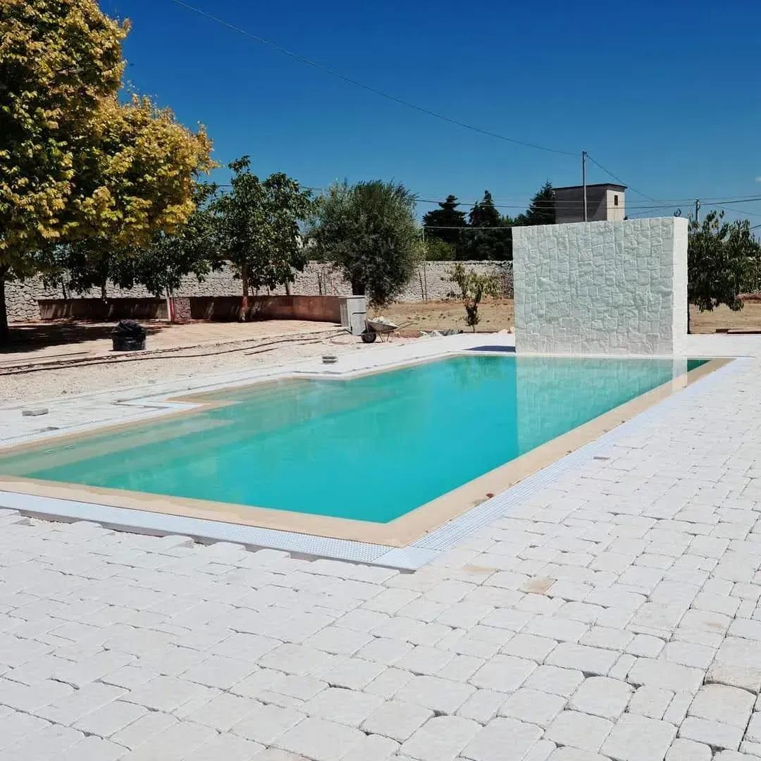 Piscina con acqua turchese, terrazza piastrellata bianca e una parete bianca testurizzata sotto un cielo azzurro.