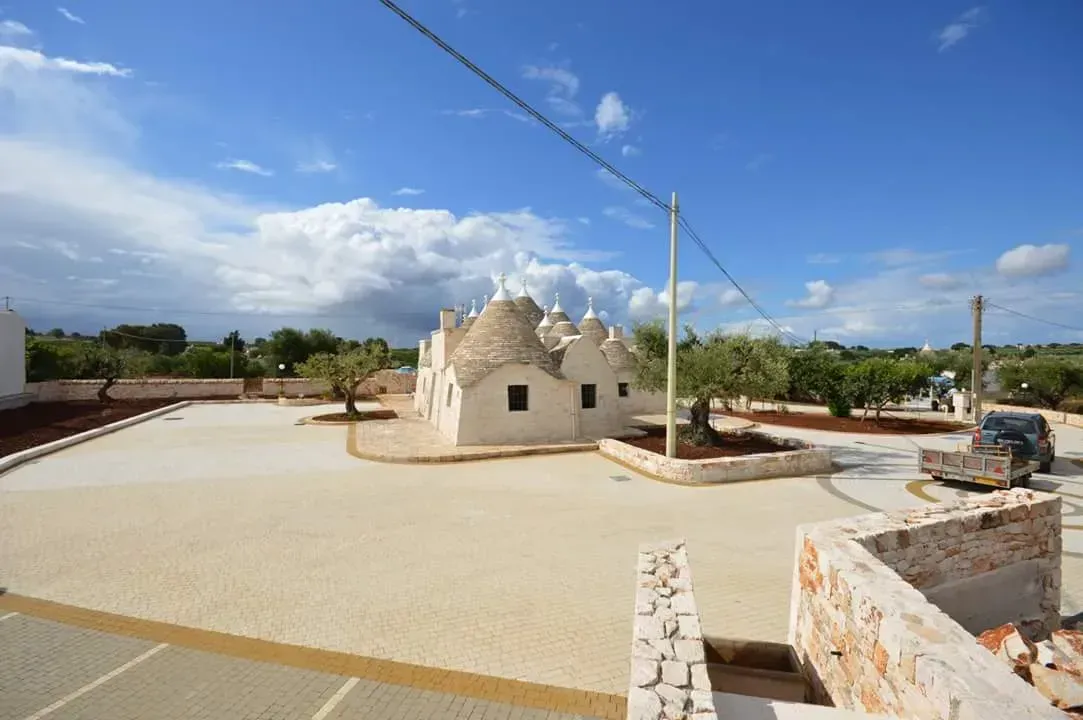 Trulli imbiancati a calce con tetti conici, ulivi e un cortile chiaro sotto un cielo azzurro.