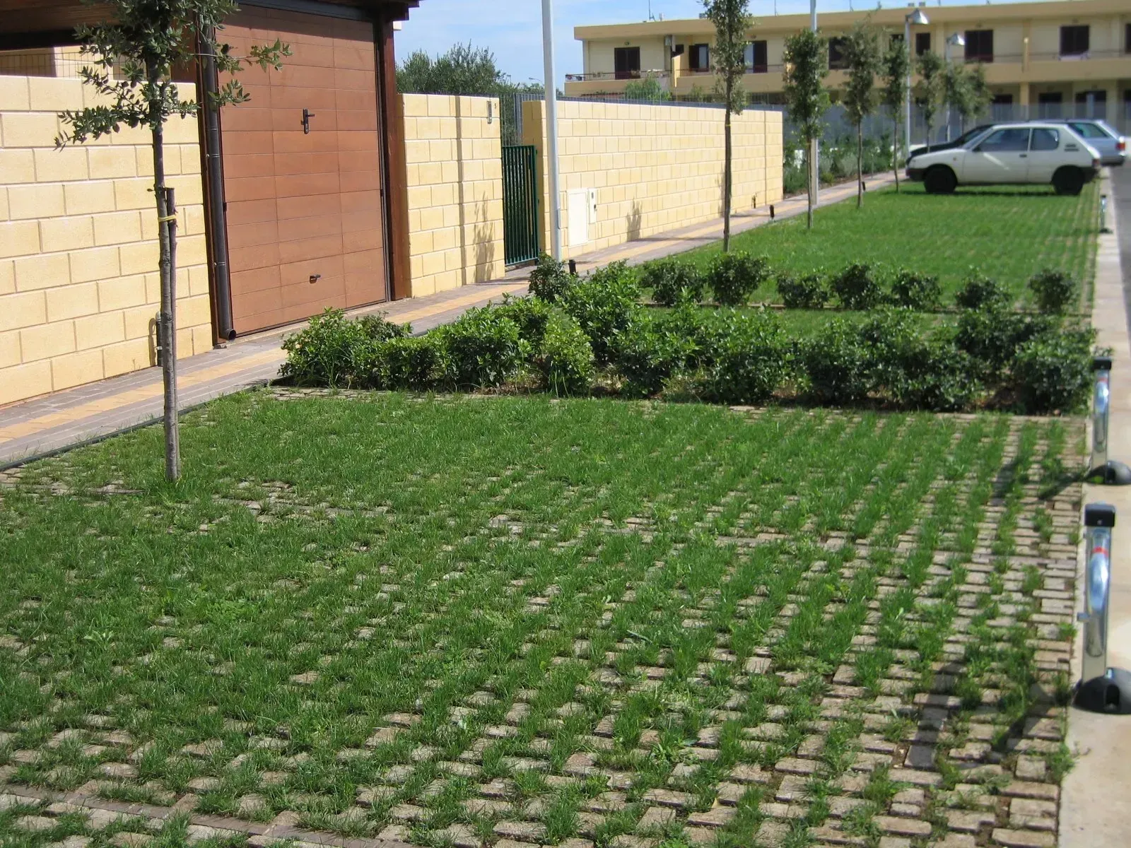 Area di parcheggio con prato verde all'interno di pavimentazioni in cemento, delimitata da siepi e da un edificio in mattoni.