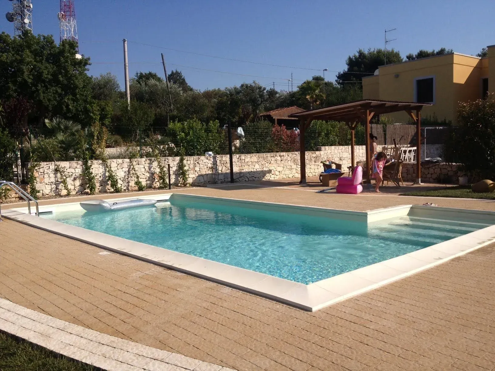 Piscina con acqua azzurra e patio circostante. Bambini che giocano nelle vicinanze. Pergolato in legno. Edificio giallo sullo sfondo.