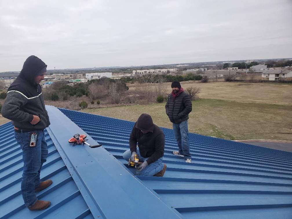 Three workers on a blue metal roof, one kneeling with tools, overlooking a grassy area.