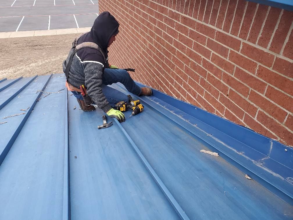 Worker kneeling on a blue metal roof beside a brick wall, using tools to fix a seam