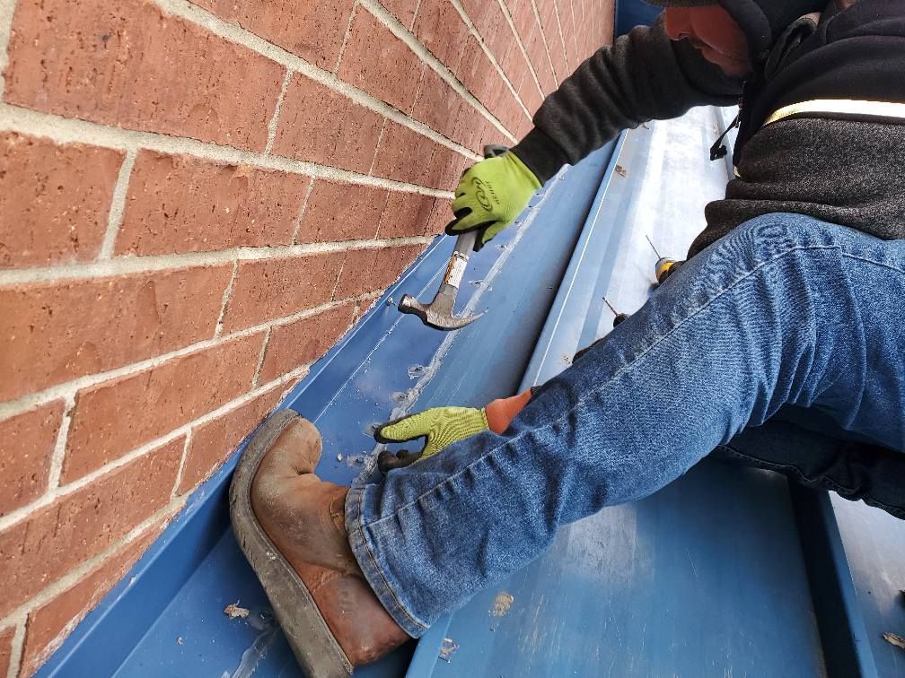 Workers installing blue roofing panels along a brick wall, wearing gloves and boots.