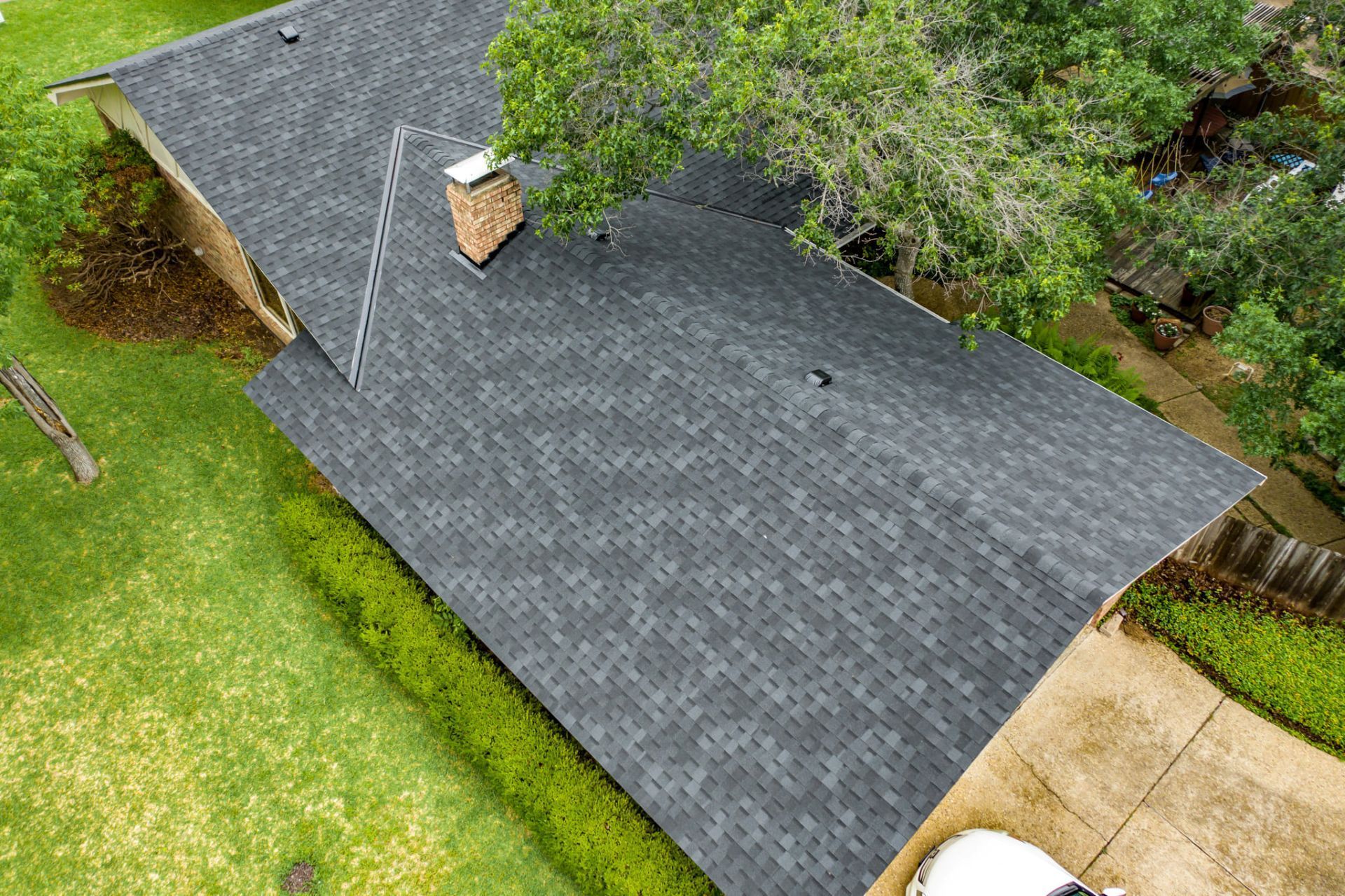 Aerial view of a house roof with gray shingles, a brick chimney, and surrounding green lawn and trees