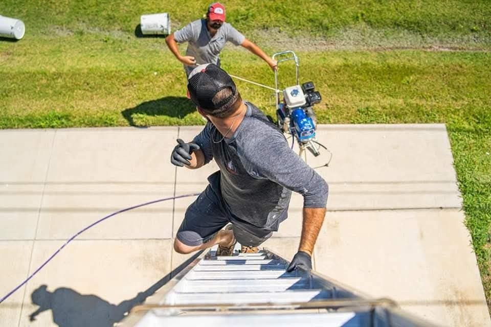 Worker climbing a ladder while another person steadies it with a rope on a grassy roof edge