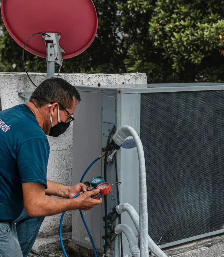 Man in mask working on AC unit on a rooftop, satellite dish in background.