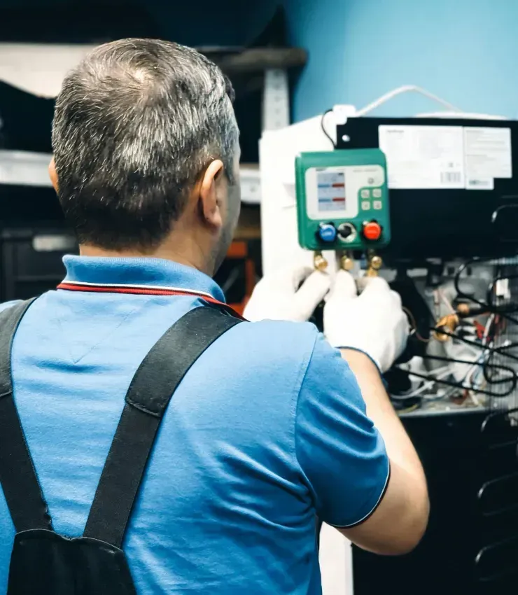 Mechanic in blue shirt and overalls working on a machine, wearing gloves.