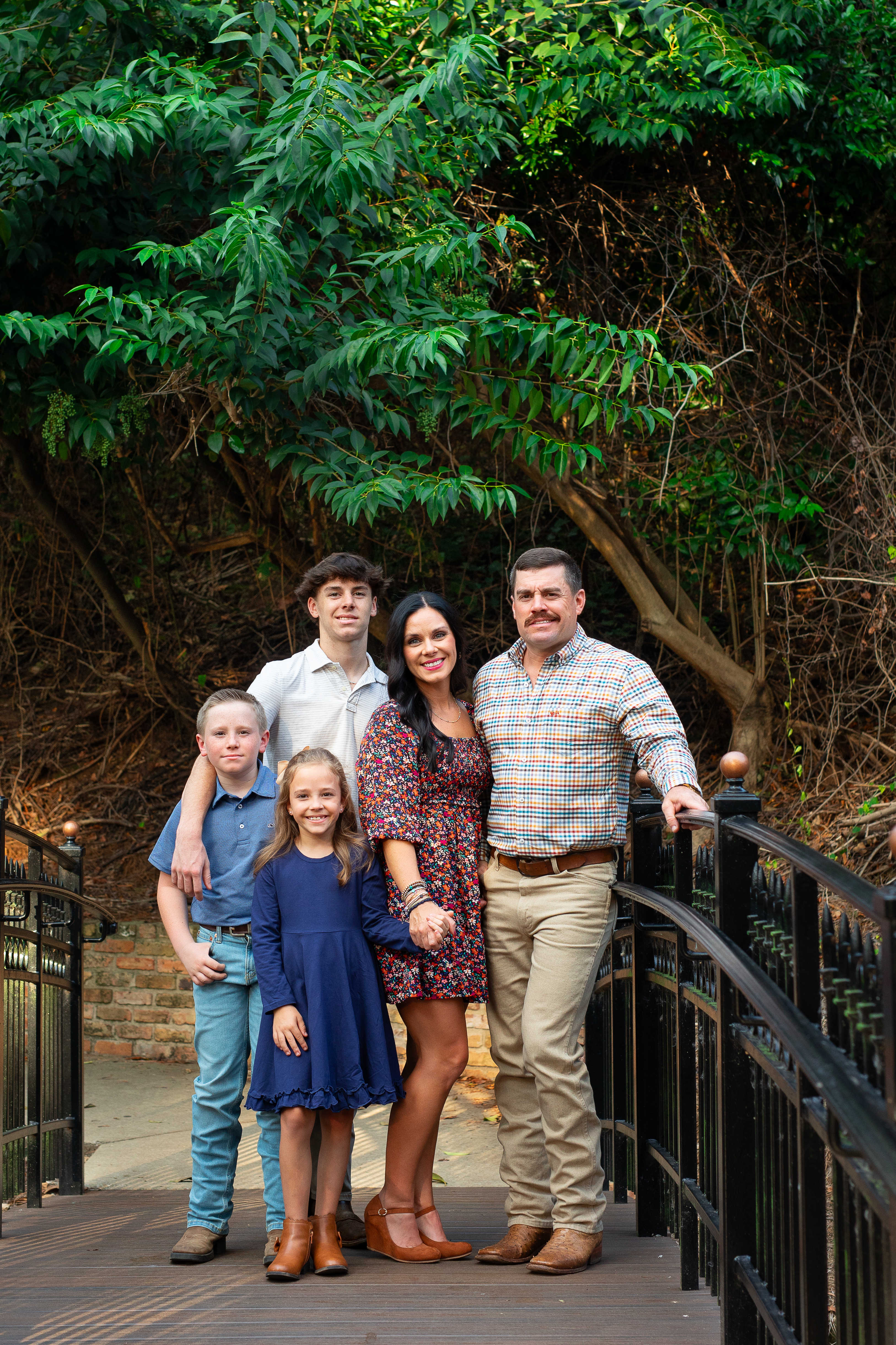 Family sitting on grass smiling; boy in vest, girl with bow, man, woman, teen boy.