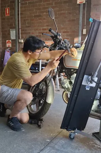 Person kneeling beside a motorcycle, working on its front end dent repair in a garage-like space